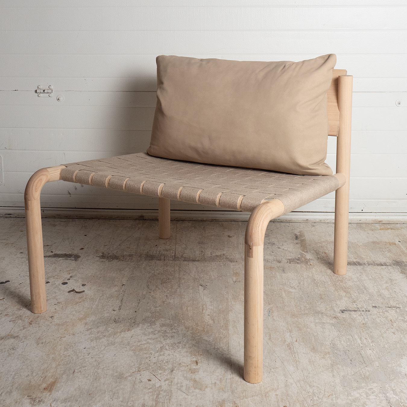 Ash lounge chair with a  leather cushion  and linen seat on a concrete floor against a white wall.
